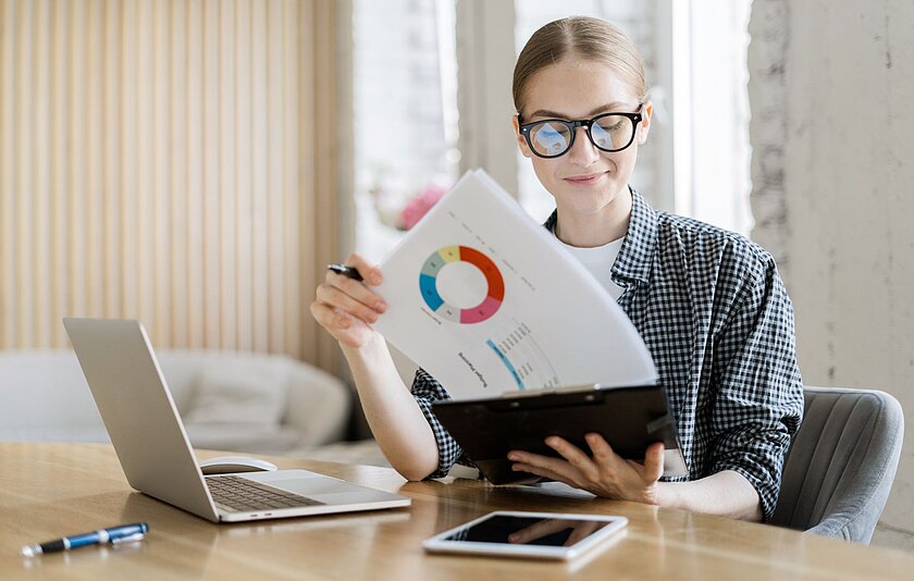Junge Frau mit schwarzer Brille sitzt an einem Schreibtisch. Sie hält ein Klemmbrett mit Statistiken in der Hand. Vor ihr stehen ein Laptop und ein Tablet.
