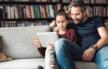 Ein Vater sitzt neben seiner Tochter auf einem Sofa. Die Tochter hält ein Tablet in der Hand und gestikuliert.