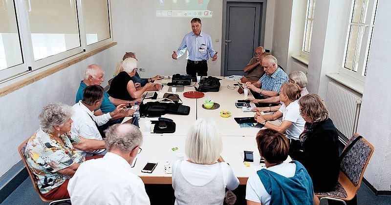 Man sieht eine Gruppe von Senioren, die mit digitalen Endgeräten an einem Tisch sitzt und auf einer Fortbildung des Senioren-Medienmentoren-Programms ist. Im Hintergrund steht ein Referent, der die Fortbildung leitet.
