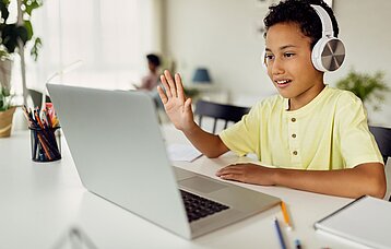 Junge mit Kopfhörern winkt bei einer Videokonferenz in den Laptop