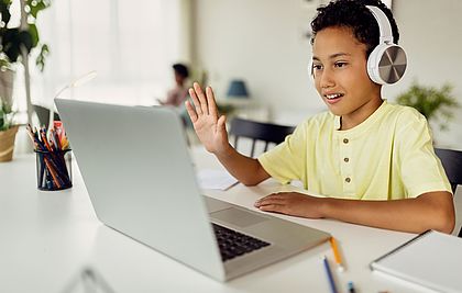 Junge mit Kopfhörern winkt bei einer Videokonferenz in den Laptop