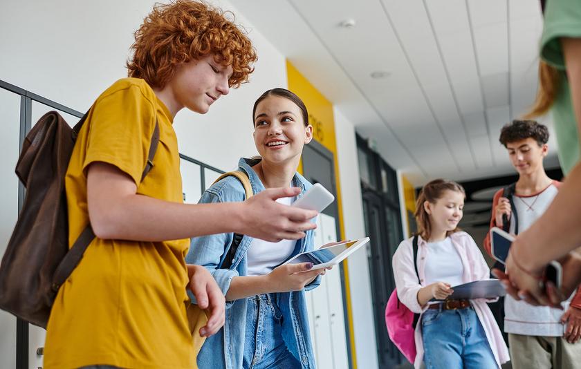 Mehrere Jugendliche stehen auf dem Flur einer Schule und unterhalten sich lachend. Alle haben Tablets oder Smartphones in der Hand.
