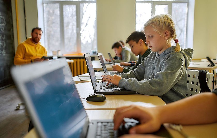 Schulklasse mit Laptops. Lehrer sitzt im Hintergrund und dbeobachtet die Schülerinnen und Schüler.