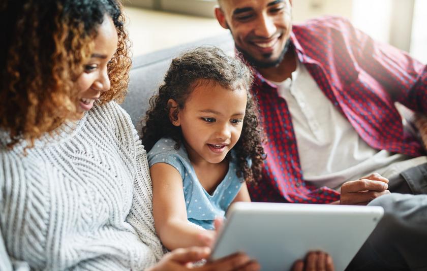 Eltern-Medienmentoren-Programm Ein Kind sitzt zwischen seiner Mutter und seinem Vater auf dem Sofa. Alle schauen gemeinsam in ein Tablet.