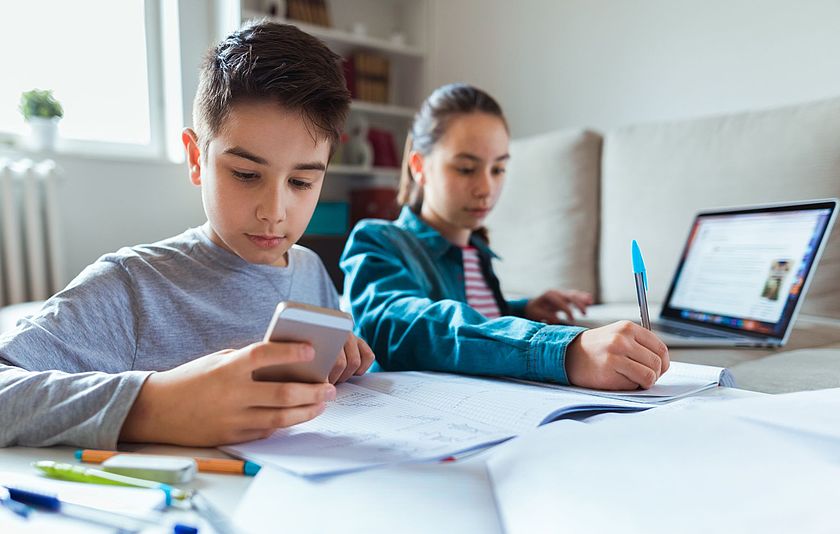 Zwei Kinder beim Hausaufgaben machen mit Smartphone und Laptop