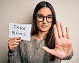 Eine junge Frau mit Brille streckt ihre Handfläche frontal nach vorne und hält in der anderen Hand einen Zettel mit der Aufschrift "Fake News".
