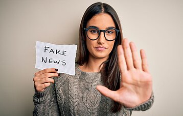 Eine junge Frau mit Brille streckt ihre Handfläche frontal nach vorne und hält in der anderen Hand einen Zettel mit der Aufschrift "Fake News".