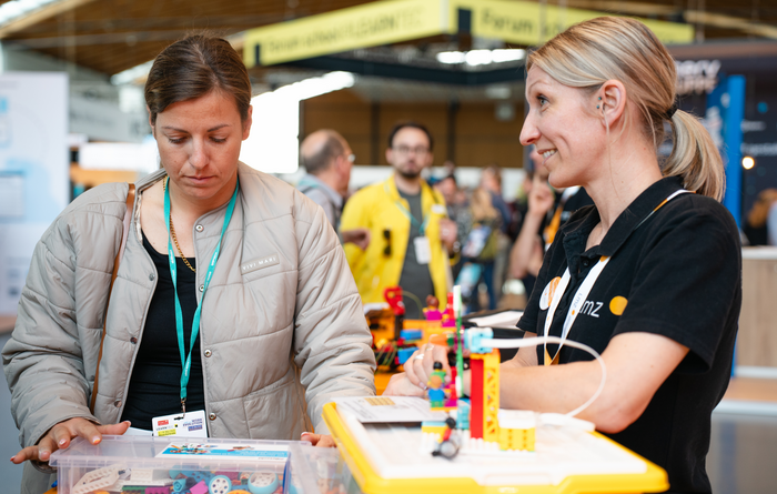 Frau im schwarzen LMZ-Polo-T-Shirt zeigt einer interessierten Messebesucherin ein Robotikset