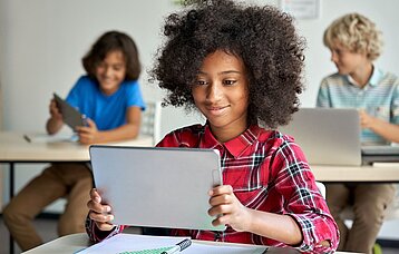 Ein Mädchen sitzt in einem Klassenzimmer und hält lächelnd ein Tablet in beiden Händen. 