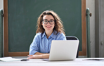 Lehrerin lächelnd vor einer Tafel am Laptop sitzend.
