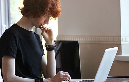 Ein Junge mit roten Haaren sieht mit nachdenklichem Gesicht auf seinen Laptop
