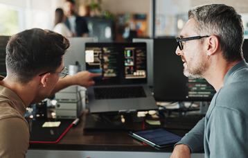 Ein Mann erklärt seinem Kollegen einen technischen Sachverhalt vor dem Rechner. Sie wirken wie Softwareentwickler beim Brainstormen oder der Arbeit. 