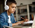Ein Junge mit Kopfhörern sitzt vor einem Laptop und macht sich auf einem Papierblock Notizen