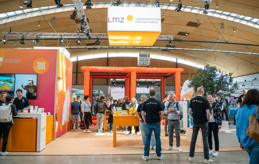 LMZ-Messestand in orangeroten Farbtönen mit Mitarbeitenden und Besuchenden