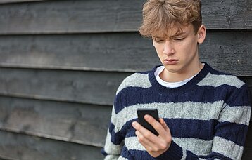 Ein Teenager-Junge in schwarz-grau gestreiftem Pullover starrt mit traurigem Blick auf sein Smartphone.