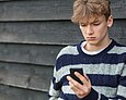 Ein Teenager-Junge in schwarz-grau gestreiftem Pullover starrt mit traurigem Blick auf sein Smartphone.
