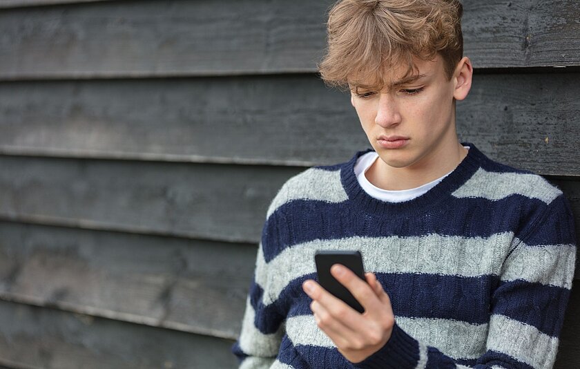 Ein Teenager-Junge in schwarz-grau gestreiftem Pullover starrt mit traurigem Blick auf sein Smartphone.