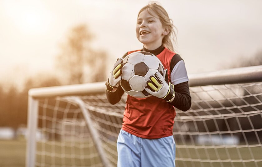 Ein Mädchen hält mit großen Torwart-Handschuhen freudestrahlend einen Fußball vor einem Fußballtor in der Hand.