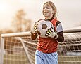 Ein Mädchen hält mit großen Torwart-Handschuhen freudestrahlend einen Fußball vor einem Fußballtor in der Hand.