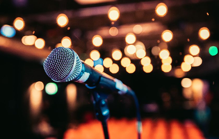 Mikrofon im Vordergrund mit Scheinwerfern und roten Sitzplätzen im Hintergrund.
