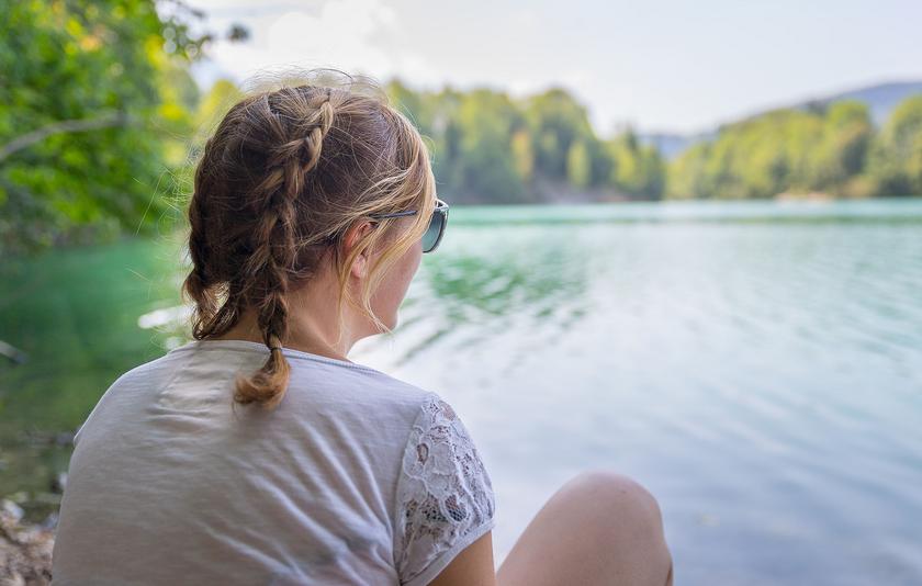 Blonde, junge Frau mit Zöpfen sitzt am Ufer und schaut auf einen ruhigen See. Im Hintergrund erscheinen grüne Wälder.