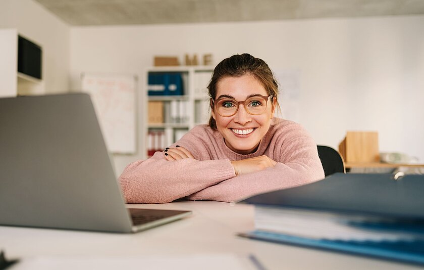 Junge Frau mit Brille lächelt in die Kamera und stützt sich mit verschränkten Armen auf einem Schreibtisch mit Laptop und Ordner ab. 