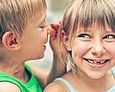Kleiner Junge flüstert lachendem Mädchen mit Zahnlücke etwas ins Ohr