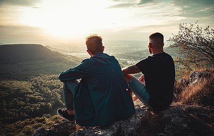 Zwei Freunde sitzen am Trauf eines Berges und schauen bei Sonnenuntergang ins Tal. Ihre Blicke schweifen in die Ferne.