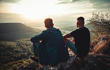 Zwei Freunde sitzen am Trauf eines Berges und schauen bei Sonnenuntergang ins Tal. Ihre Blicke schweifen in die Ferne.