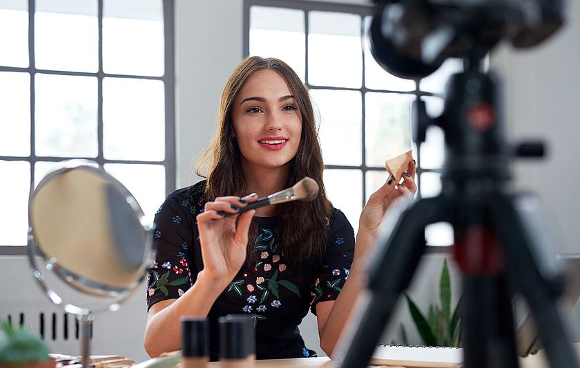 Junge Frau sitzt vor einer Kamera und hält Kosmetik-Artikel davor. 
