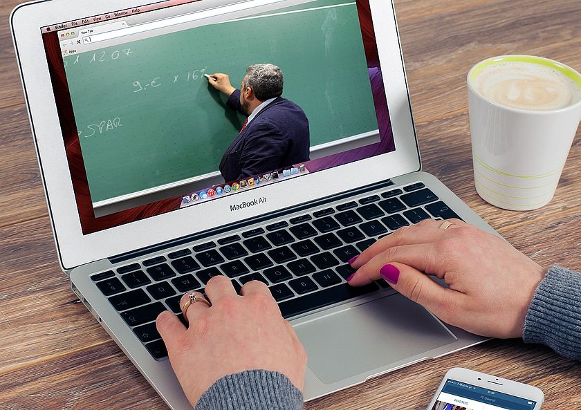 Hände auf einer Laptop-Tastatur, dessen Display einen Lehrer zeigt vor einer Schultafel. Hände auf einer Laptop-Tastatur, dessen Display einen Lehrer zeigt vor einer Schultafel.