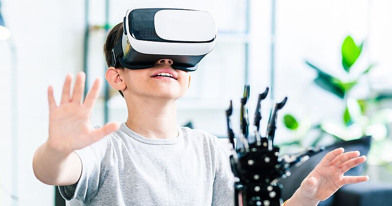 Ein Junge hat eine VR-Brille auf und hält seine Hände in die Luft. Vor ihm steht eine Roboterhand.