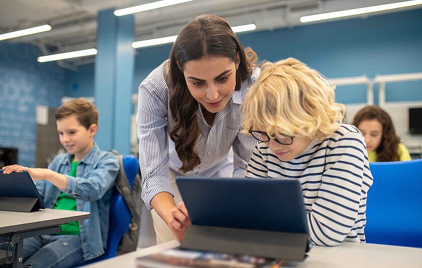 Lehrerin beugt sich über einen Schüler mit Brille und blondem lockogen Haar, um ihm am Tablet eteas zu erkläeren.