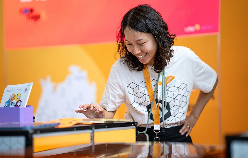 Makermobil Mitarbeiterin Lien Ha Duong Viet Eine Frau mit halblangen dunklen Haaren steht vor dem lasercutter Modul des makermobils des Landesmedienzentrums. Sie arbeitet an dem Modul und laechelt.