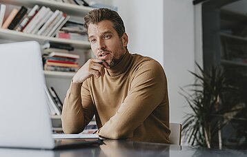 Ein Mann mittleren Alters schaut nachdenklich auf einen Laptop und hat eine Hand an sein Kinn gelegt. In der Hand hält er einen Stift.