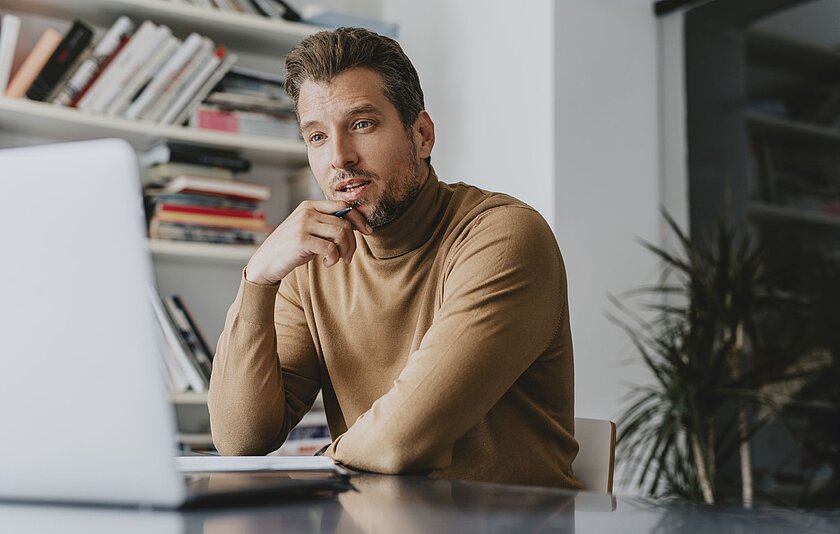 Ein Mann mittleren Alters schaut nachdenklich auf einen Laptop und hat eine Hand an sein Kinn gelegt. In der Hand hält er einen Stift.