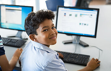 Grundschüler sitzt vor seinem Computer in der Klasse und dreht sich lächelnd zur Kamera um.